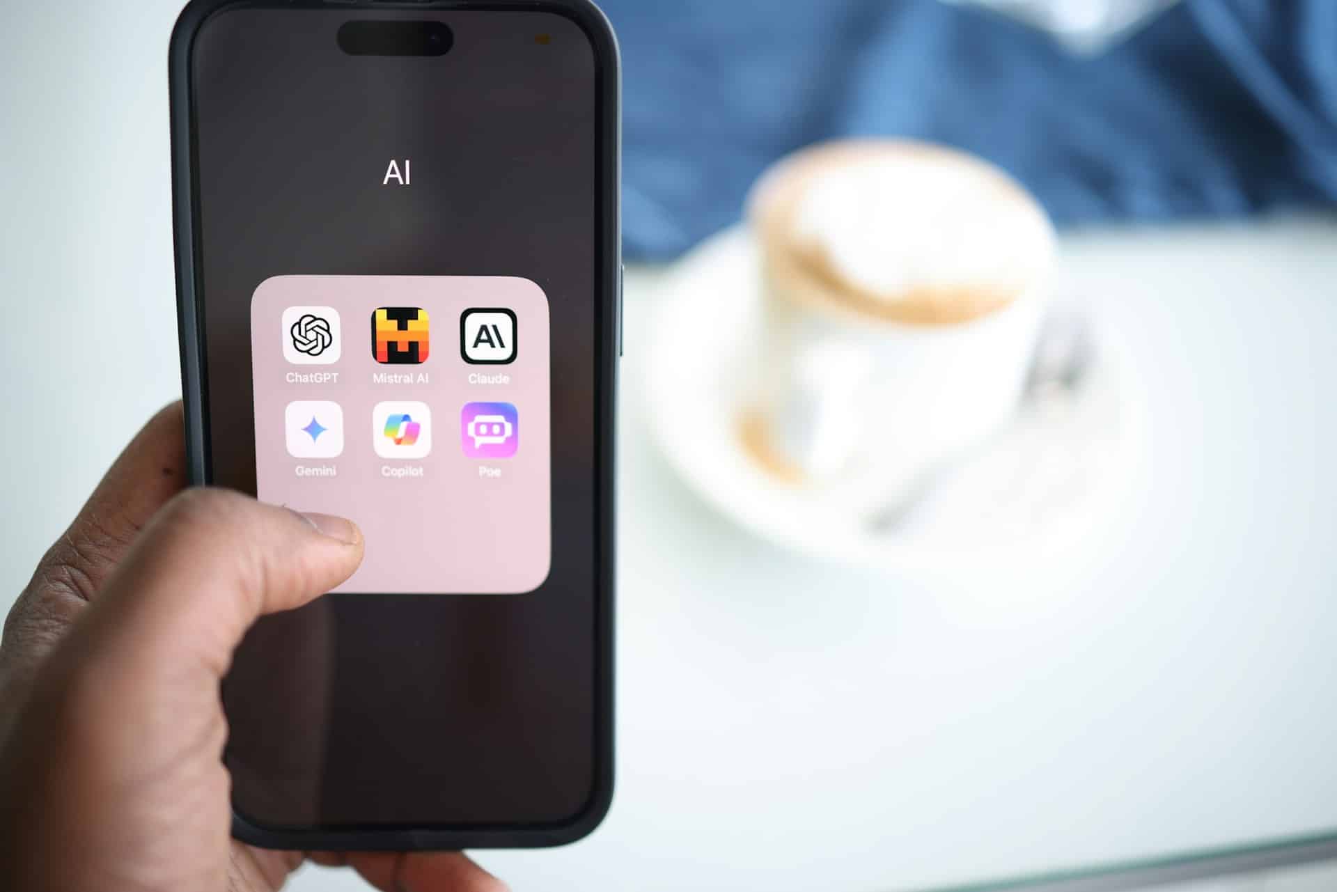Une main tient un smartphone dans lequel se trouve un dossier intitulé AI, avec des icônes d'applications telles que ChatGPT et Copilot - des outils parfaits pour l'optimisation des moteurs de recherche et l'optimisation générative des moteurs. En arrière-plan flou, une tasse de café attend sur sa soucoupe.