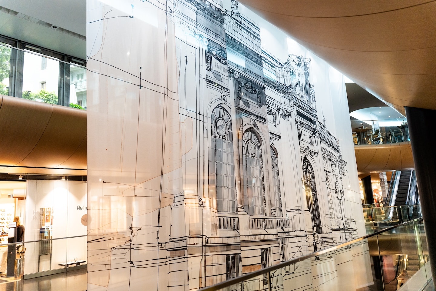 Un grand panneau semi-transparent présentant un dessin architectural du musée d'art et d'histoire (MAH) de Genève est exposé à l'intérieur d'un centre commercial moderne avec des garde-corps en verre et des plafonds incurvés.